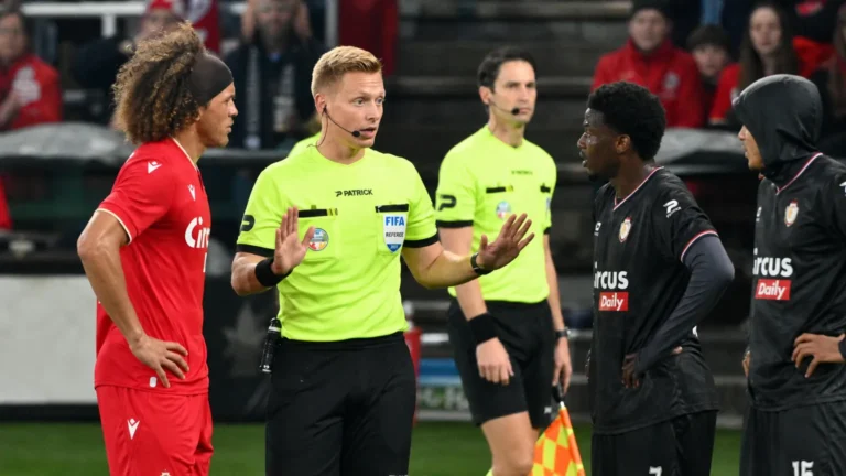 Een scheidsrechter spreekt met spelers in rode en donkere tenues tijdens een voetbalwedstrijd.