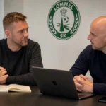 Twee medewerkers overleggen in een vergaderruimte van Lommel SK met een clubembleem aan de muur.