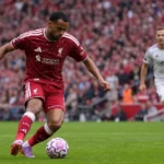 Een Liverpool-speler in donkerrood tenue schiet de bal tijdens een wedstrijd op Anfield.