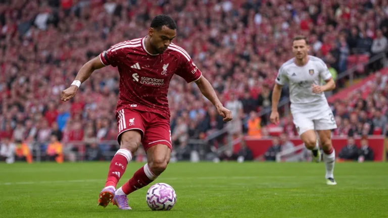 Een Liverpool-speler in donkerrood tenue schiet de bal tijdens een wedstrijd op Anfield.