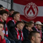 Supporters van KV Kortrijk in een tribune met een clubbanner tijdens een avondwedstrijd.