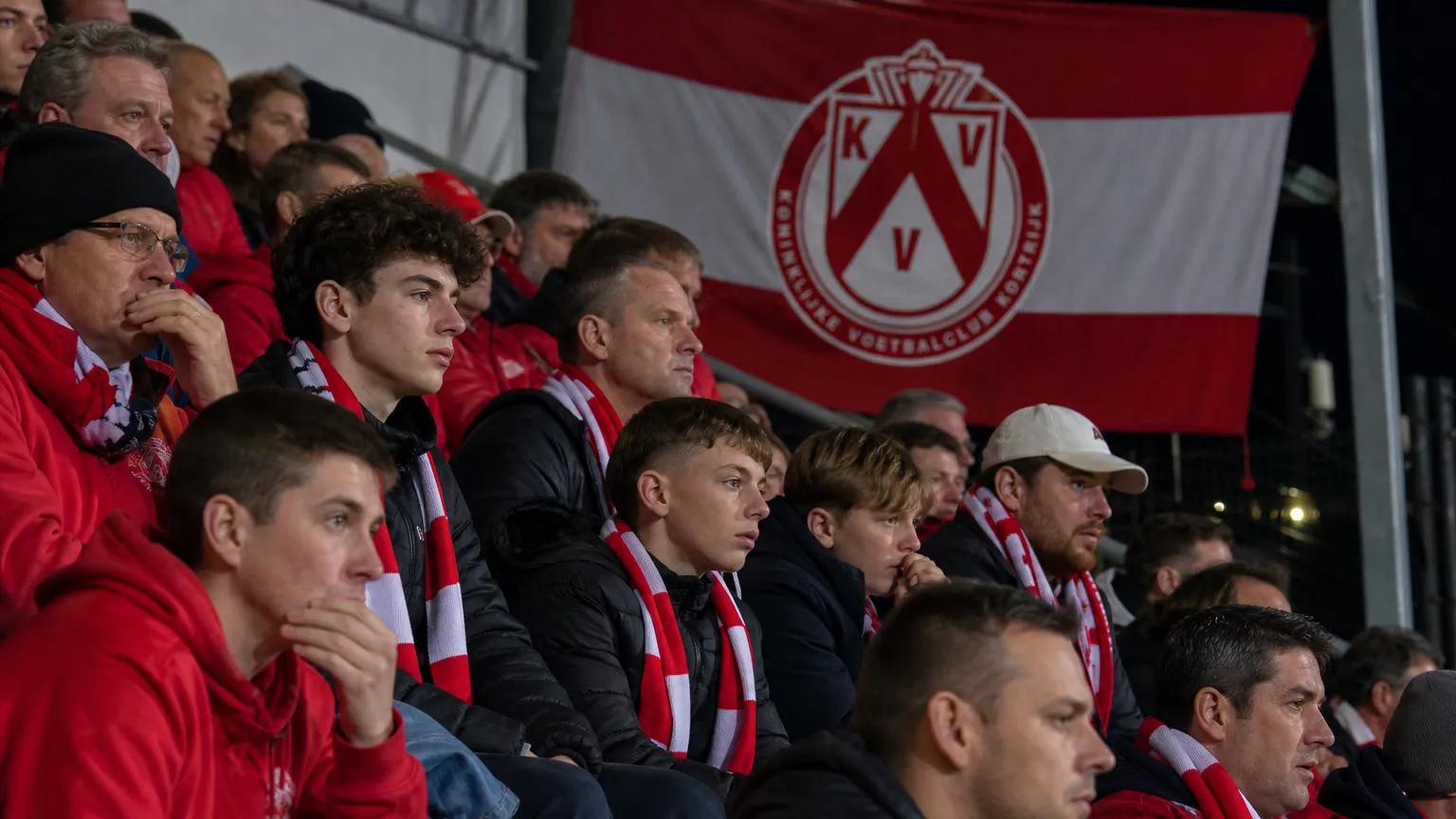 Supporters van KV Kortrijk in een tribune met een clubbanner tijdens een avondwedstrijd.