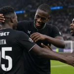 Drie spelers in zwarte wedstrijdtenues vieren samen op het veld.