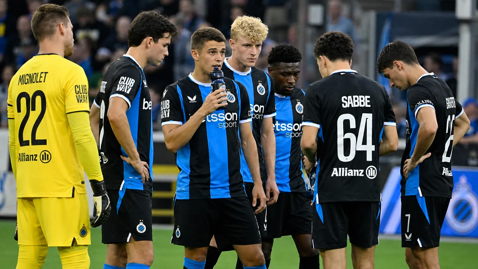 Een groep voetballers in zwart-blauwe tenues tijdens een spelonderbreking naast het veld.