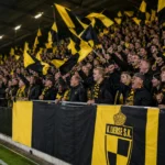 Lierse-supporters zwaaien met geel-zwarte vlaggen in een voetbalstadion.