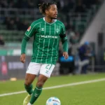 Samuel Mbangula in een groen gestreept Werder Bremen-shirt aan de rand van het veld.