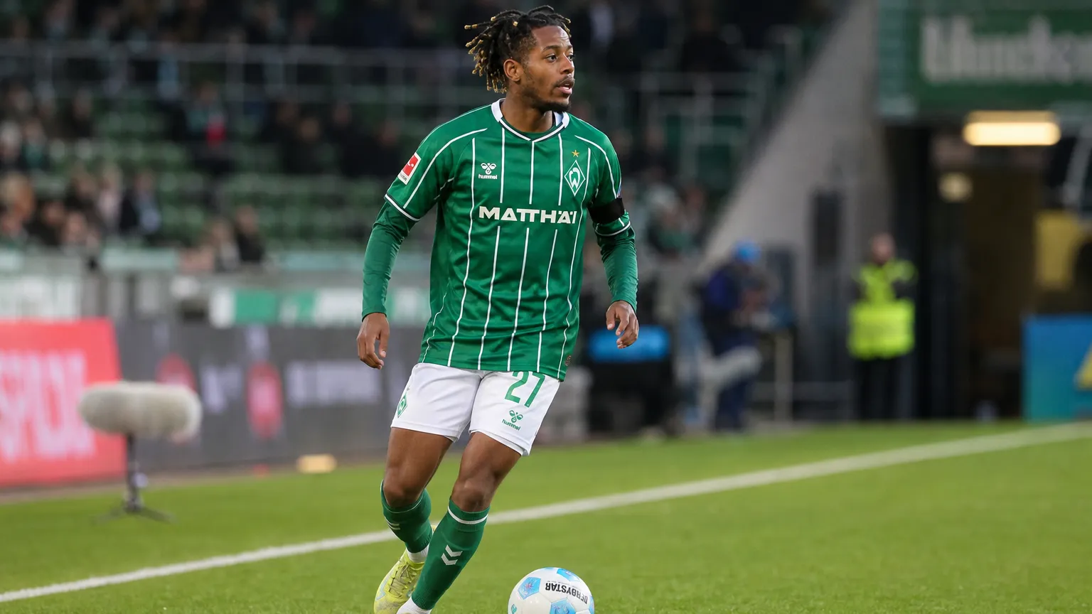 Samuel Mbangula in een groen gestreept Werder Bremen-shirt aan de rand van het veld.