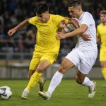 Niet-identificeerbare voetballers in gele en witte tenues vechten om de bal op een verlicht veld.