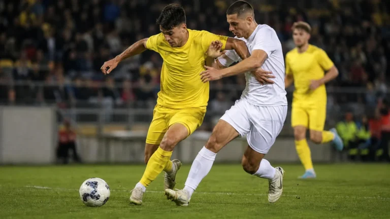 Niet-identificeerbare voetballers in gele en witte tenues vechten om de bal op een verlicht veld.