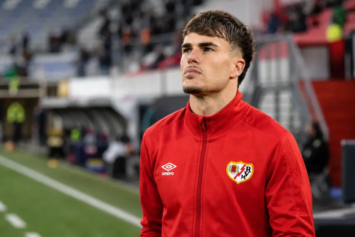 Pep Chavarria in een rode trainingsjas langs de zijlijn van een stadion.