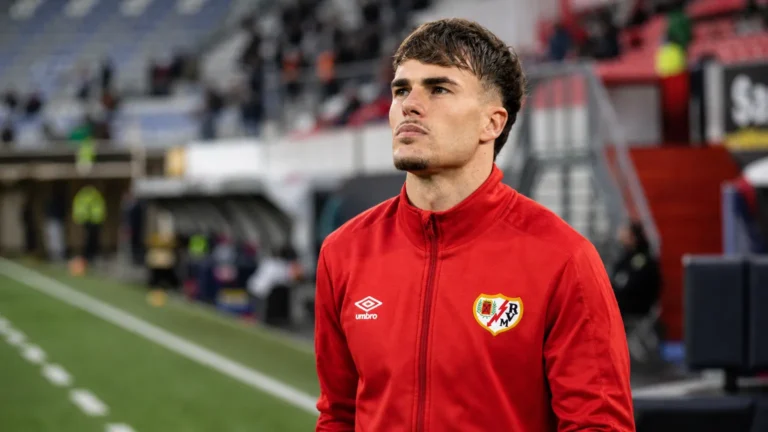 Pep Chavarria in een rode trainingsjas langs de zijlijn van een stadion.