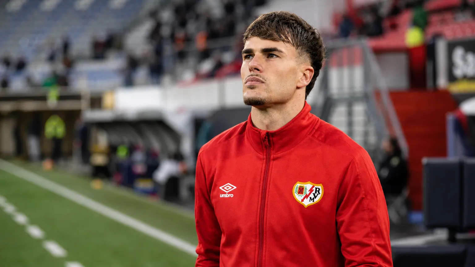 Pep Chavarria in een rode trainingsjas langs de zijlijn van een stadion.