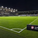 Een verlicht voetbalveld met een Jupiler Pro League-bord langs de zijlijn.