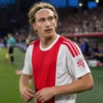 Mika Godts in een rood-wit shirt langs de zijlijn van een voetbalstadion.