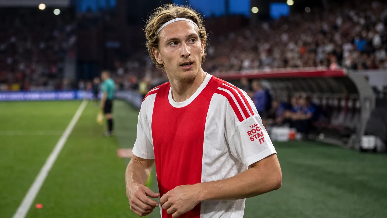 Mika Godts in een rood-wit shirt langs de zijlijn van een voetbalstadion.