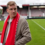 Michiel Jonckheere staat met een rode sjaal aan de rand van een voetbalveld.