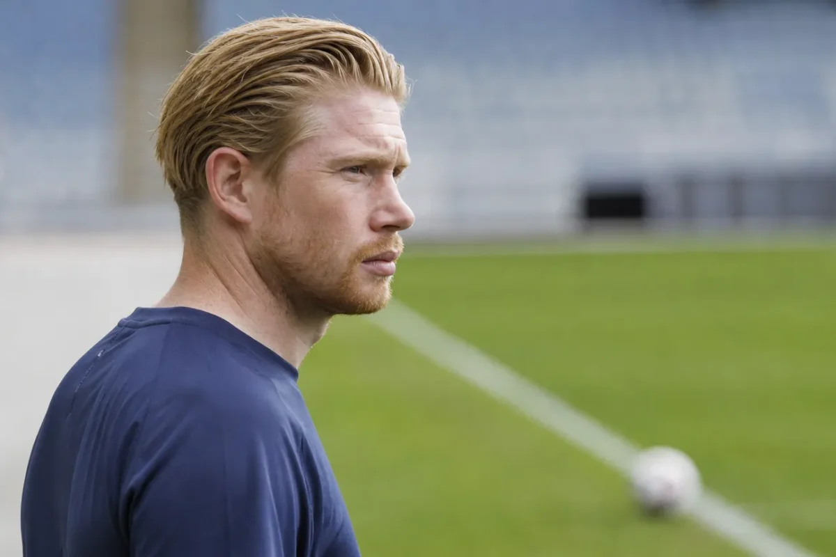 Kevin De Bruyne in neutrale sportkleding aan de rand van een voetbalveld.