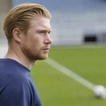 Kevin De Bruyne in neutrale sportkleding aan de rand van een voetbalveld.