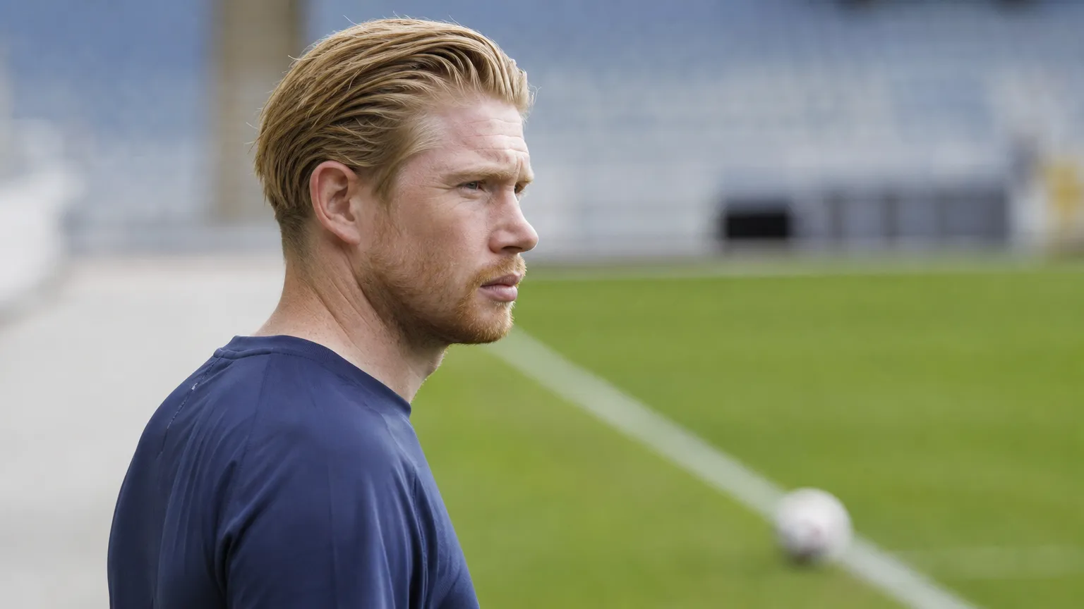 Kevin De Bruyne in neutrale sportkleding aan de rand van een voetbalveld.