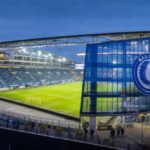 De blauw verlichte Planet Group Arena van KAA Gent met het speelveld, de overdekte tribunes en glazen gevel duidelijk zichtbaar.