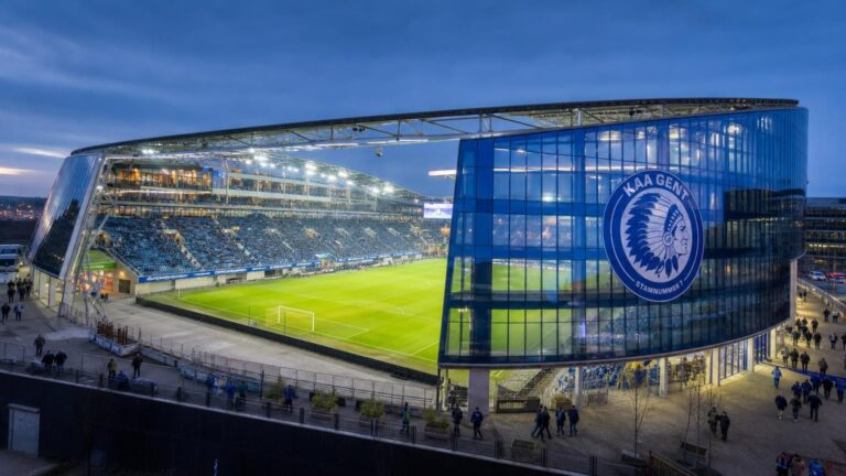 De blauw verlichte Planet Group Arena van KAA Gent met het speelveld, de overdekte tribunes en glazen gevel duidelijk zichtbaar.