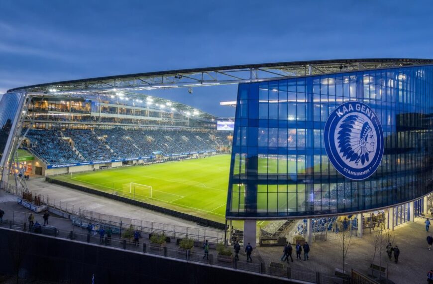 De blauw verlichte Planet Group Arena van KAA Gent met het speelveld, de overdekte tribunes en glazen gevel duidelijk zichtbaar.
