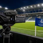 Een tv-camera langs het veld met een KAA Gent-vlag in een Zweeds voetbalstadion voor de aftrap.