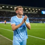 Max Dean applaudisseert langs het voetbalveld in een lichtblauw tenue.