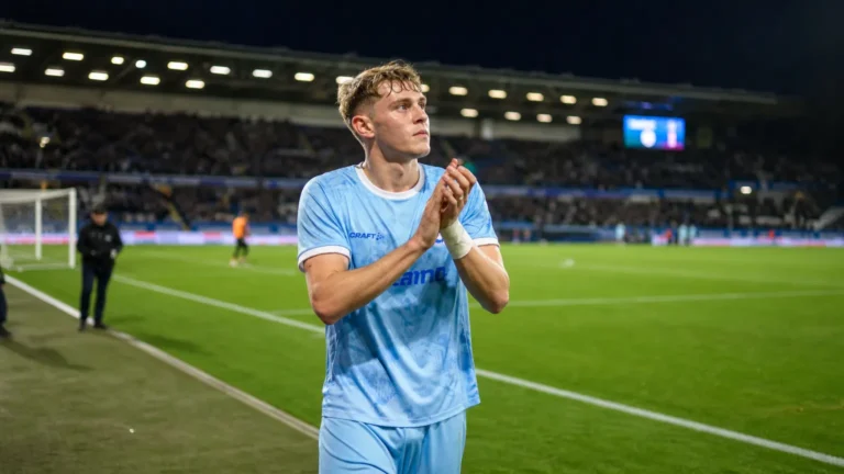 Max Dean applaudisseert langs het voetbalveld in een lichtblauw tenue.