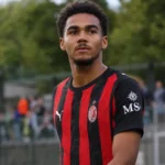 David Odogu in een zwart-rood gestreept AC Milan-tenue naast een voetbalveld.