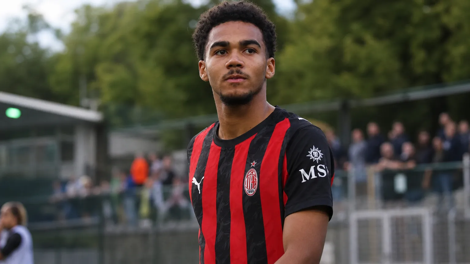 David Odogu in een zwart-rood gestreept AC Milan-tenue naast een voetbalveld.