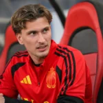 Mika Godts in een rood shirt in de dug-out van een stadion.