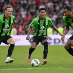 Drie voetballers in groen-zwarte tenues reageren op het veld.