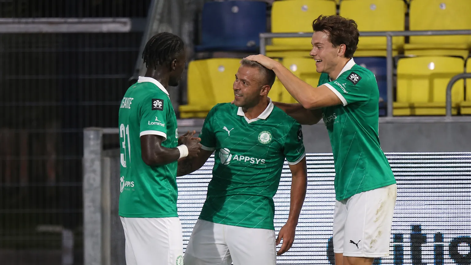 Drie spelers van Lommel SK in groene wedstrijdshirts bij de rand van het veld.