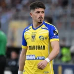 Arbnor Muja in het geel-blauwe tenue van STVV langs de zijlijn van een voetbalveld.