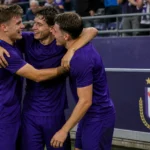Drie voetballers in paarse uitrusting vieren samen voor een discreet RSC Anderlecht-bord.