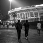 Anonieme voetballiefhebbers wandelen in de avond langs een stadionomgeving in Kampala.