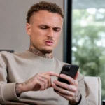 Noa Lang kijkt op zijn smartphone in een rustige binnenruimte.