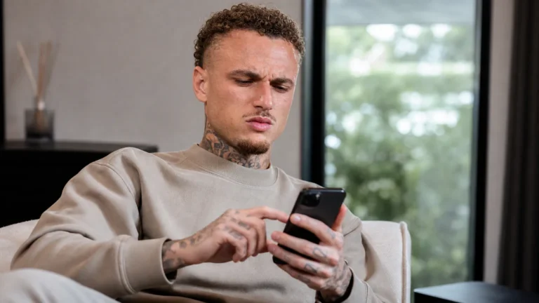Noa Lang kijkt op zijn smartphone in een rustige binnenruimte.
