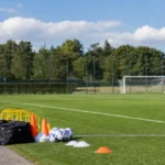 Een leeg trainingsveld met oefenmateriaal en een KAA Gent-vlag aan de rand.