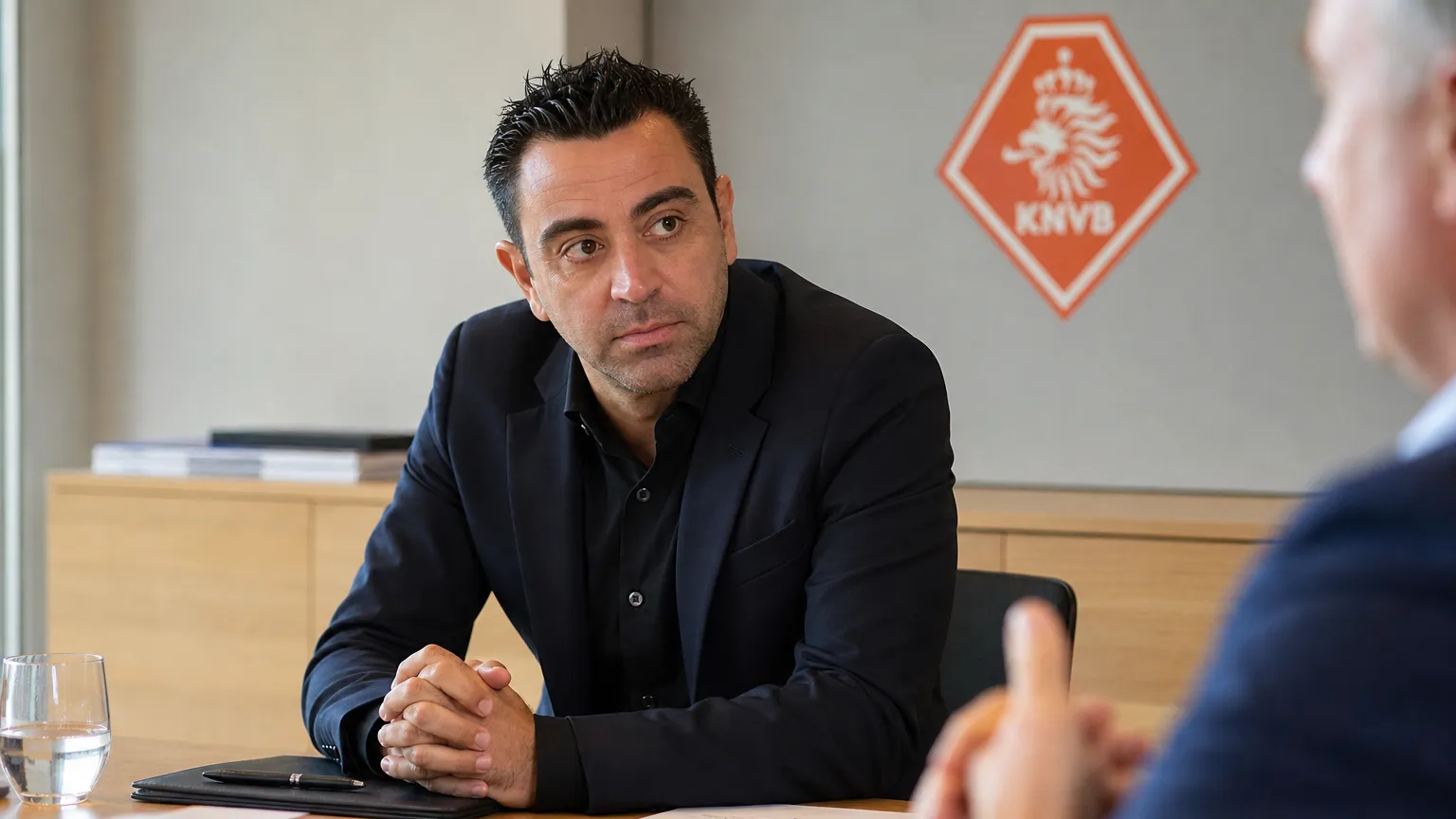 Xavi Hernández tijdens een gesprek aan een vergadertafel met een discreet KNVB-logo op de wand.