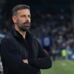 Ruud van Nistelrooij in donkere stafkleding aan de rand van een voetbalveld.