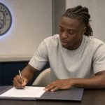 Jérémy Doku ondertekent een contract aan een tafel in een ruimte van Manchester City.