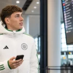 Arne Engels in een witte Celtic-jas met telefoon in een luchthavenvertrekhal.