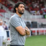 Marvin Compper staat in een grijze polo langs de zijlijn van een voetbalstadion.