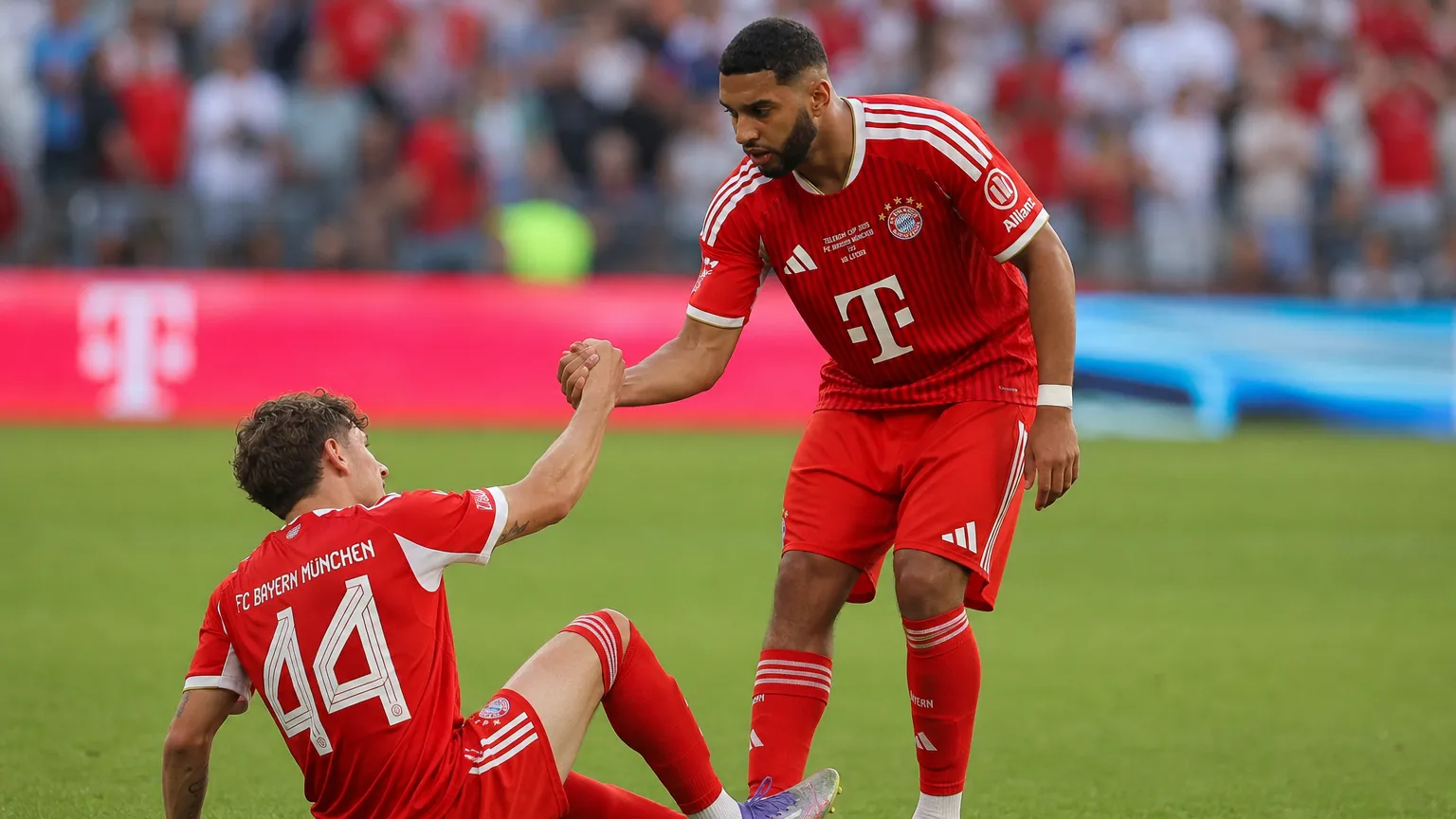 Ismael Saibari in rood Bayern-tenue helpt een zittende ploegmaat tijdens een oefenwedstrijd.