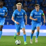 Drie spelers van KRC Genk in lichtblauwe wedstrijdtenues tijdens een competitiewedstrijd.