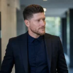 Sébastien Pocognoli in een donkere jas in een neutrale voetbalmediaomgeving.