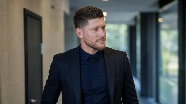 Sébastien Pocognoli in een donkere jas in een neutrale voetbalmediaomgeving.