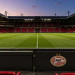 Een leeg voetbalveld in een PSV-stadion met een discreet PSV-embleem aan de rand van de tribune.